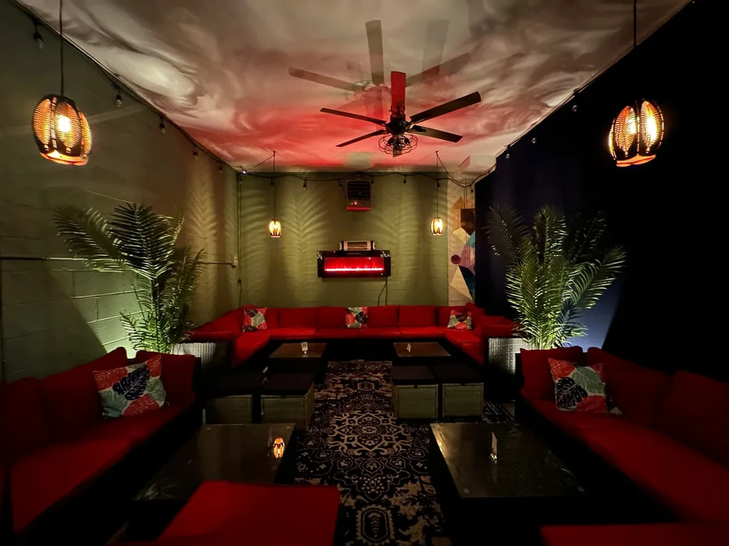 A dimly lit lounge with red couches, palm plants, and a ceiling fan.