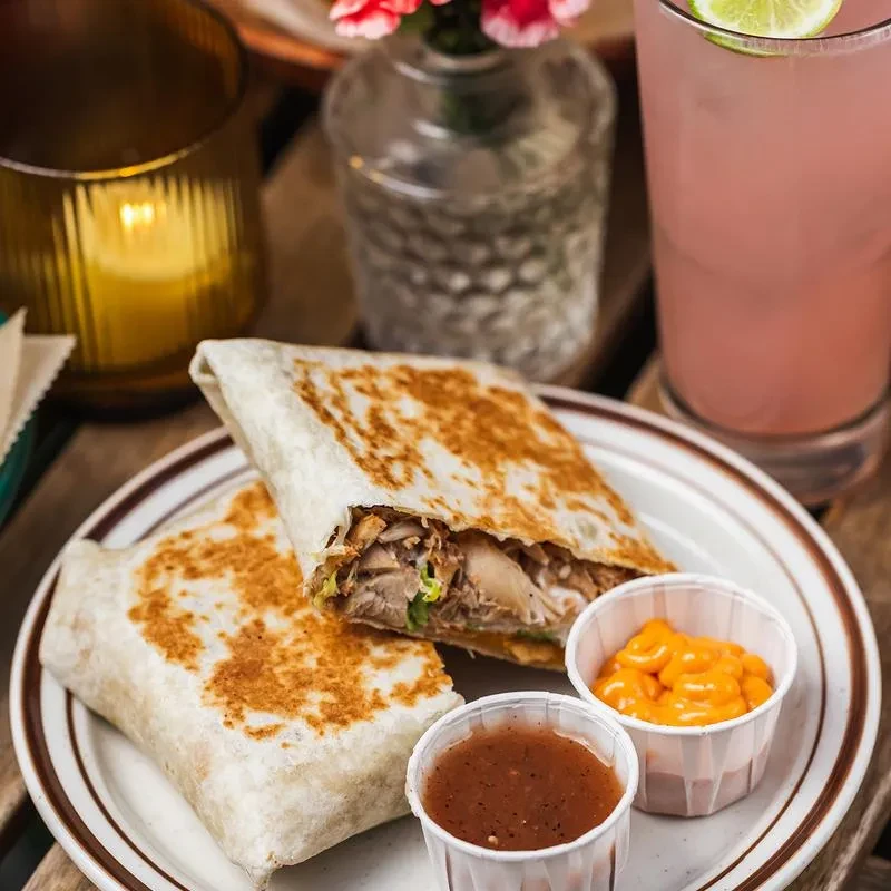 Two grilled burritos, sauce cups, pink drink, and candle on a table.