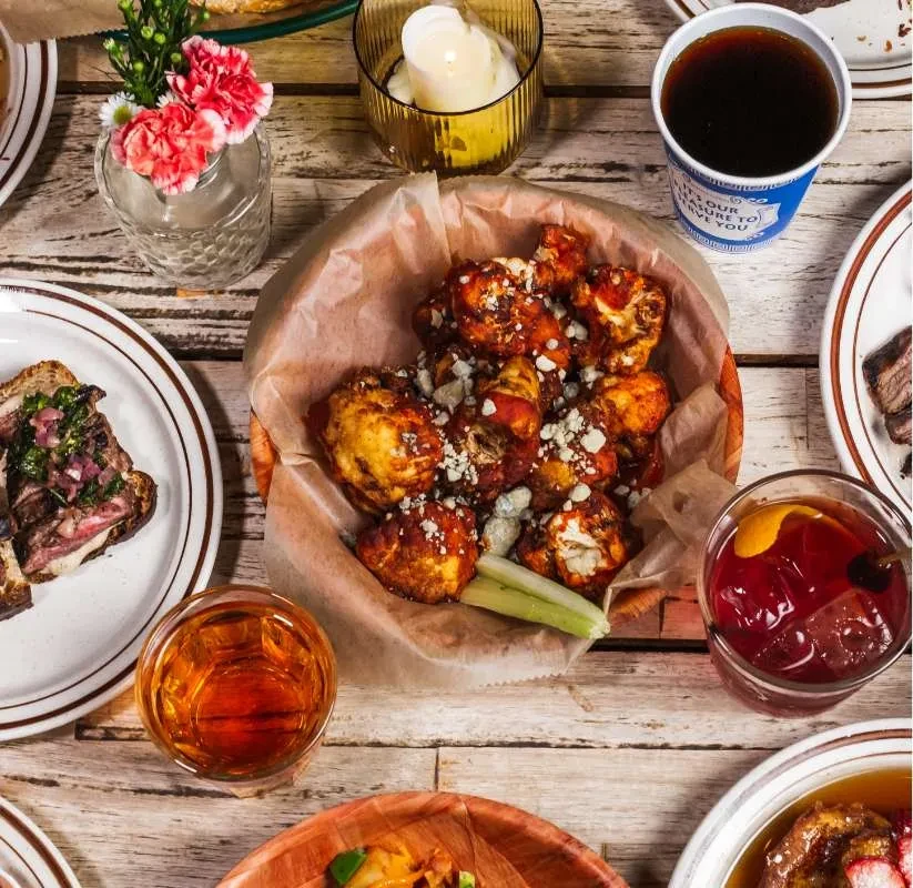 A wooden table with buffalo cauliflower, drinks, and various foods.