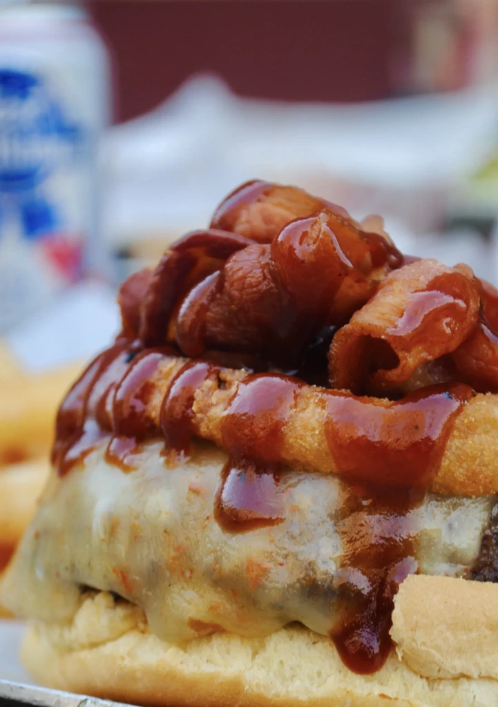 Close-up of a loaded burger with bacon, onion rings, cheese, and BBQ sauce.