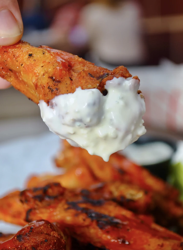 A hand dips a chicken wing into a creamy white sauce.