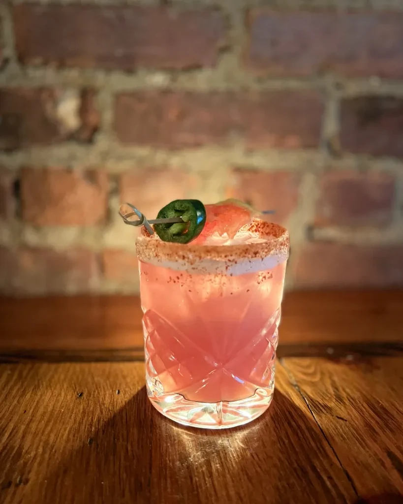 Pink cocktail with a jalapeno garnish on a wooden table.