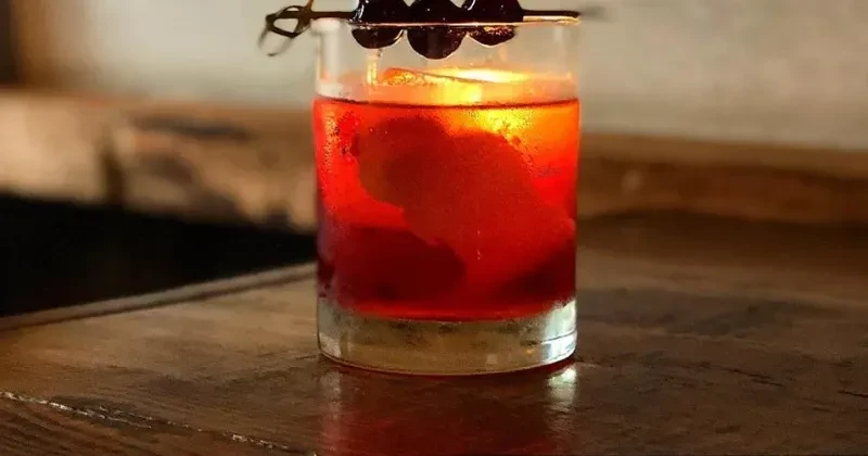 Red drink in a glass with ice, orange peel, and cherries.