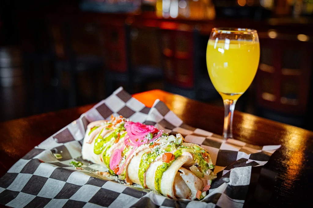Burrito with pickled onions, salsa, and green sauce next to a yellow drink.