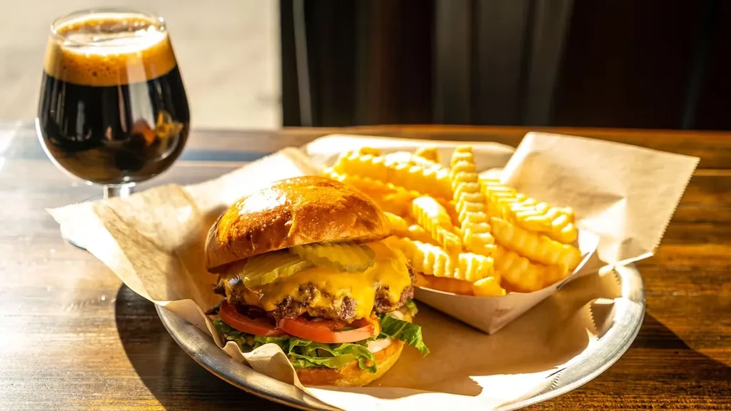 A cheeseburger with crinkle fries and a dark beer on a wooden table.