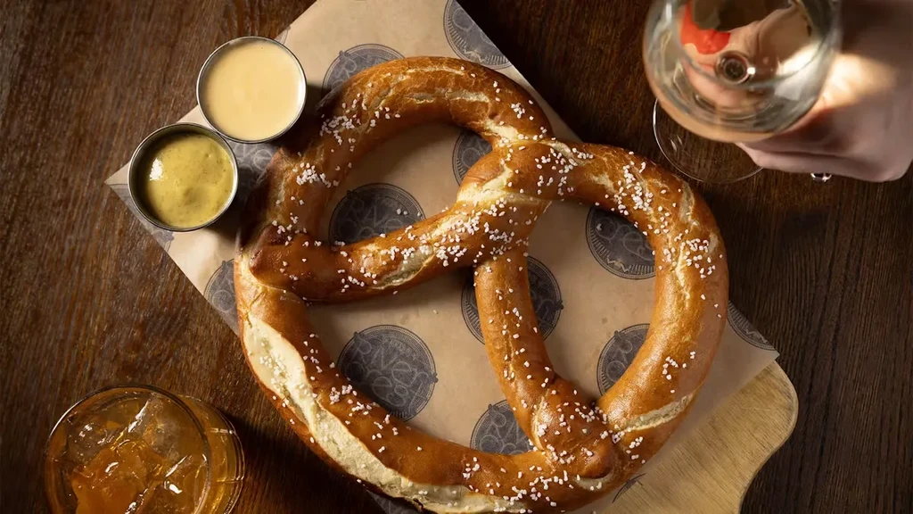 Overhead view of a pretzel with dipping sauces, drinks, and a hand.