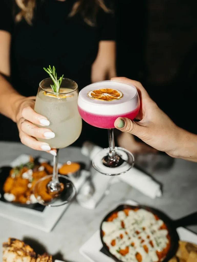 Two people toast colorful cocktails with snacks on a table.