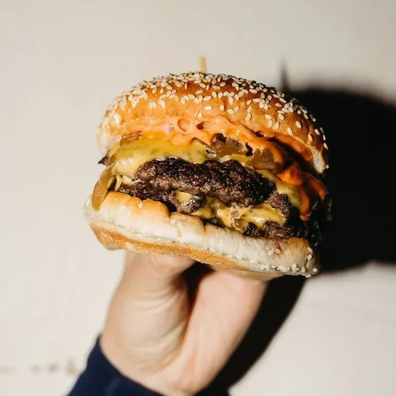 A hand holds a double cheeseburger with sesame seed bun.