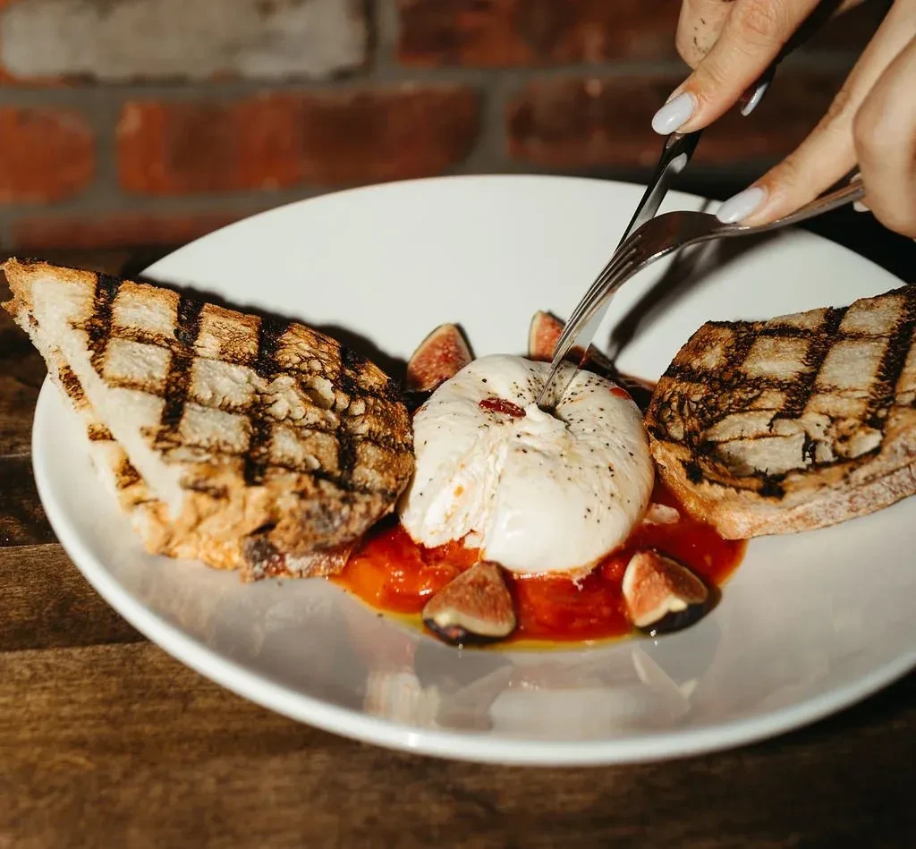 A person cutting burrata cheese, grilled bread, and figs with cutlery.