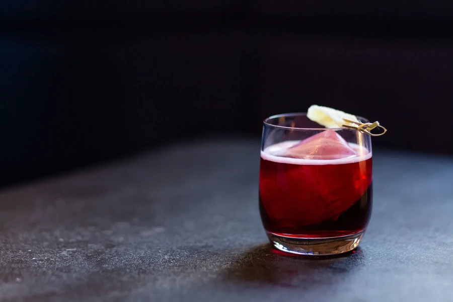 A red cocktail with a large ice cube and citrus garnish.