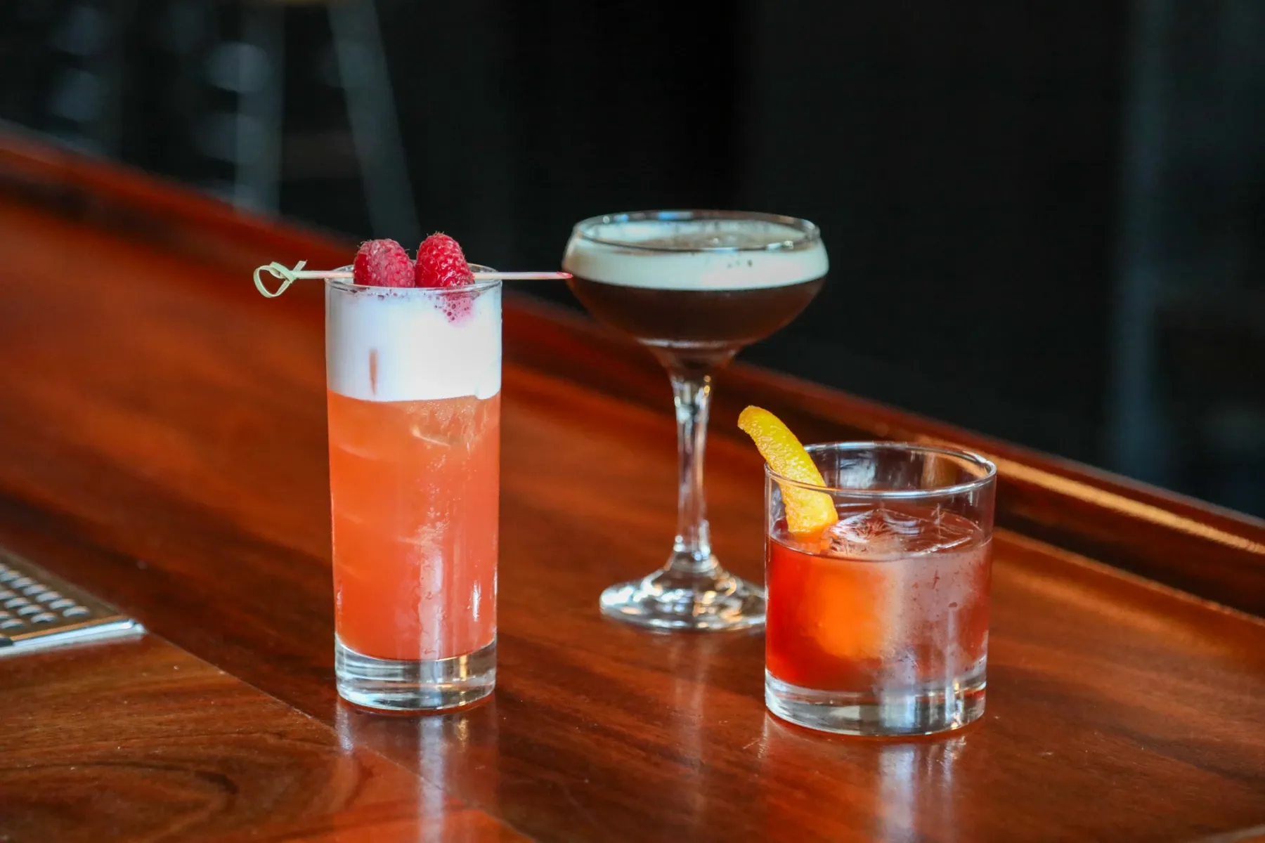 Three cocktails, two short and one tall, sit on a wooden bar.