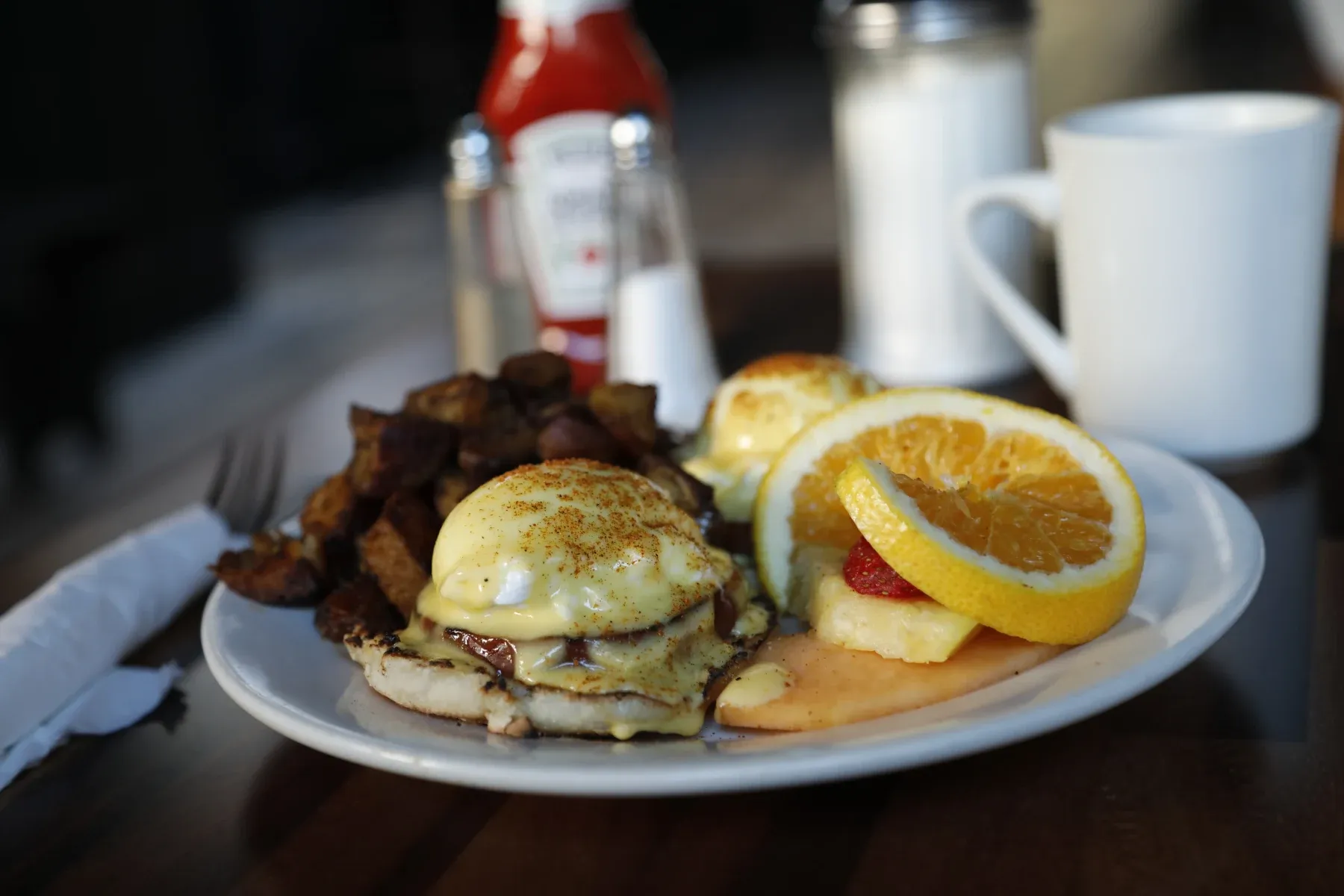Eggs Benedict, roasted potatoes, and fruit on a white plate.