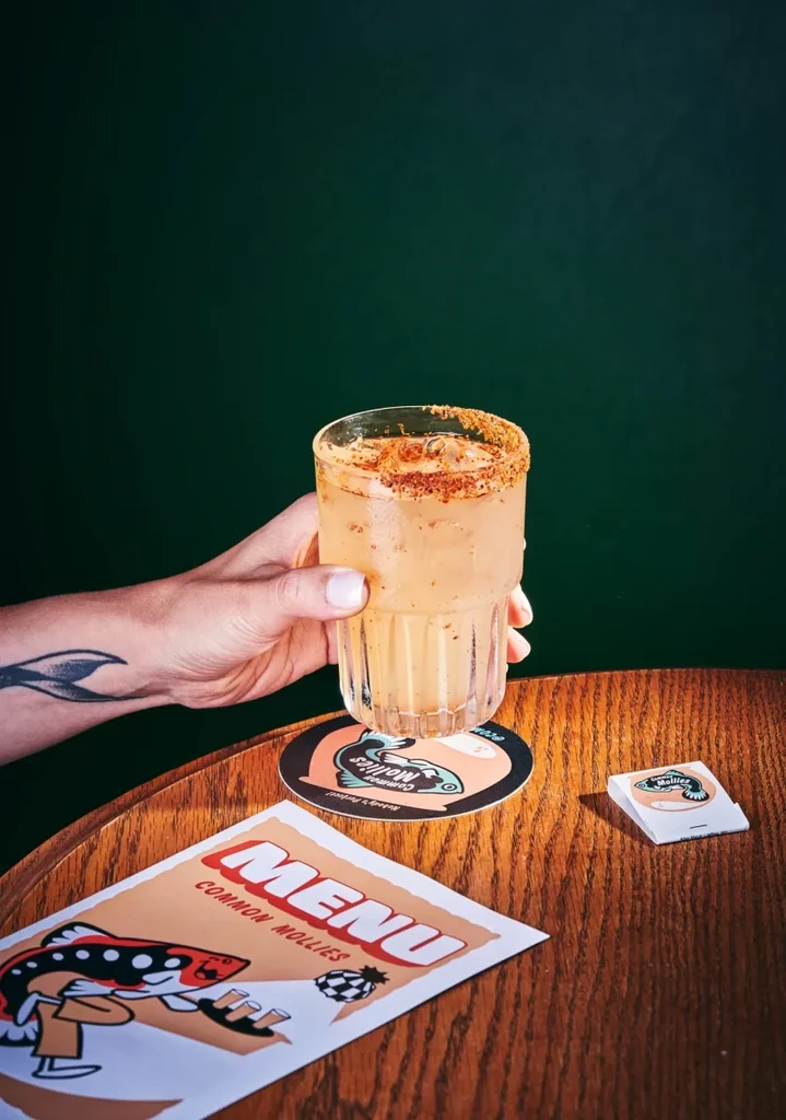 A person holds a cocktail with a spiced rim and a menu on a wooden table.