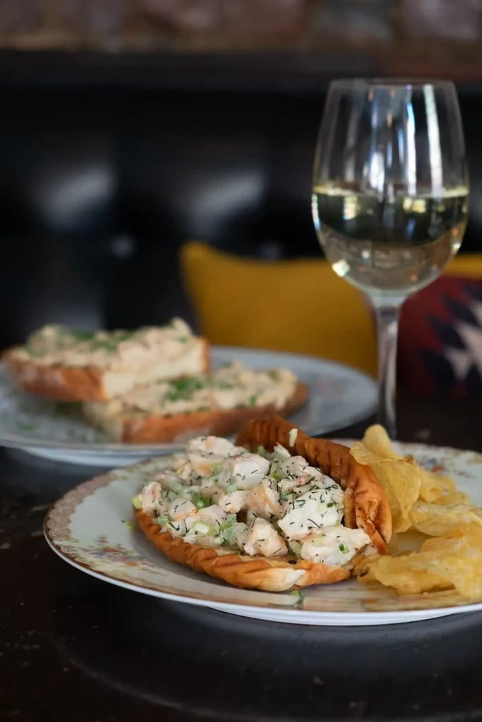 Lobster roll with potato chips, two toasts, and a glass of white wine.