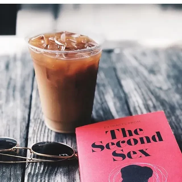 Iced coffee, sunglasses, and "The Second Sex" book on a wooden table.
