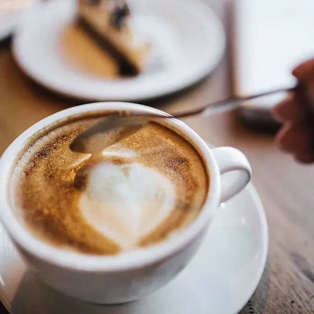 A person stirring a latte with a heart design in a white cup.