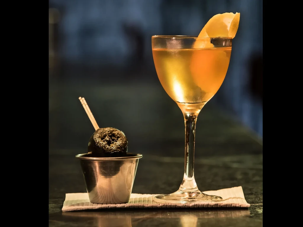A golden cocktail with a lemon twist next to a dark olive snack.