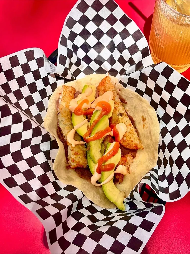Taco with hash browns, avocado, and sauce in checkered paper.
