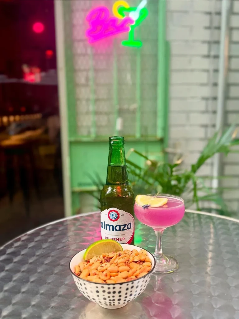 A bottle of Almaza beer, a pink cocktail, and a bowl of peanuts.
