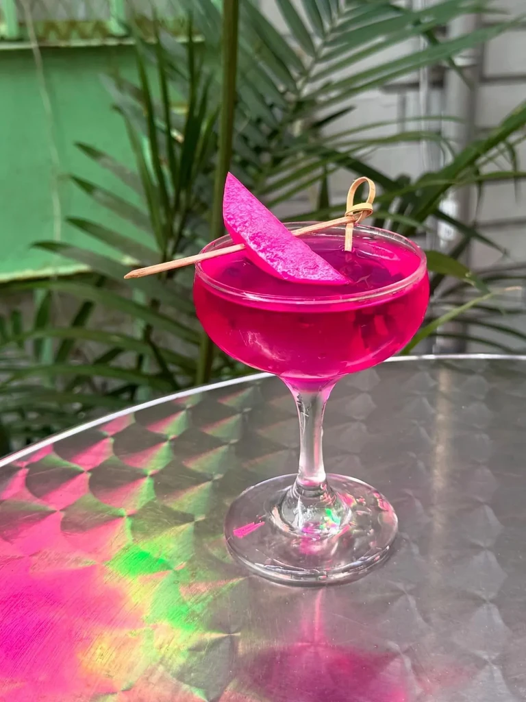 Bright pink cocktail with a fruit garnish on a reflective table.