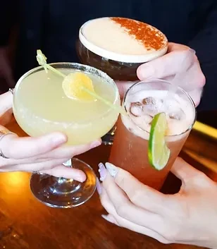 Three hands holding three different cocktails with various garnishes, toasting.