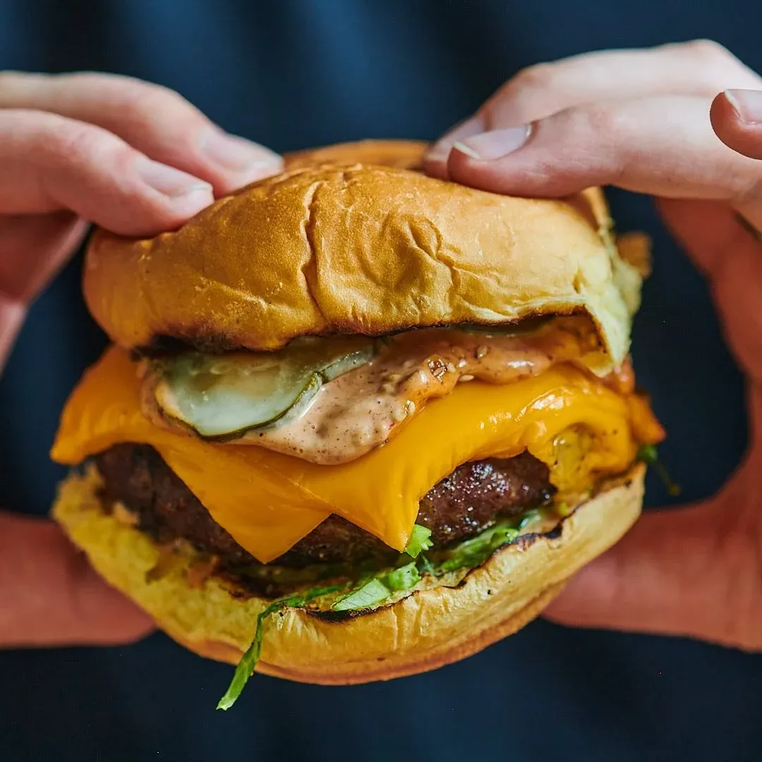 Hands hold a cheeseburger with pickles, sauce, and lettuce on a bun.