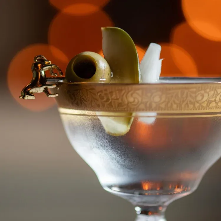 A cocktail glass with an olive, lemon peel, and a golden decorative pick.
