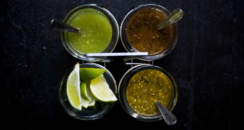 Four bowls of salsa and lime wedges on a dark background.