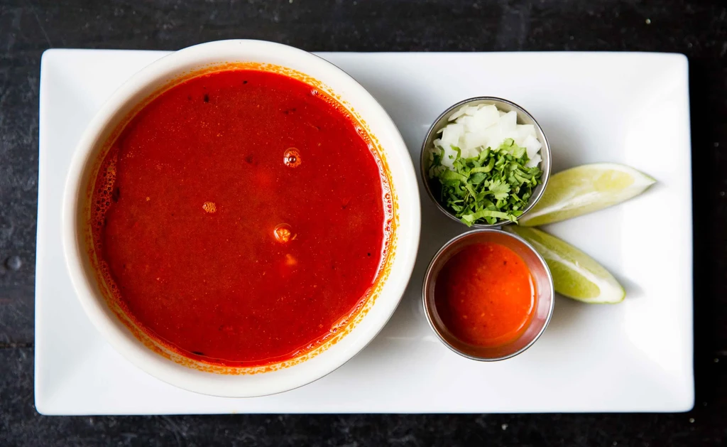 Overhead view of a red soup with sides of onion, cilantro, hot sauce, and lime.
