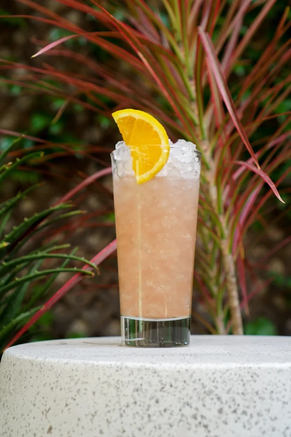 A tall glass of pink cocktail with crushed ice and an orange slice.