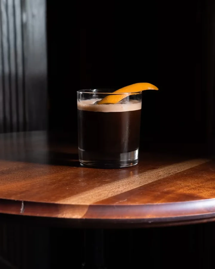 A dark drink with foam and an orange peel on a wooden table.
