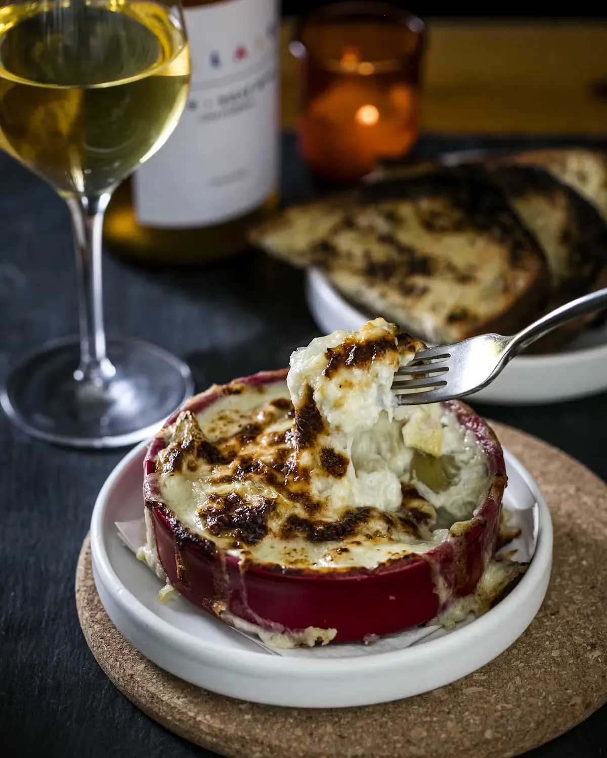 A fork lifts creamy gratin from a red ramekin with wine and toast.