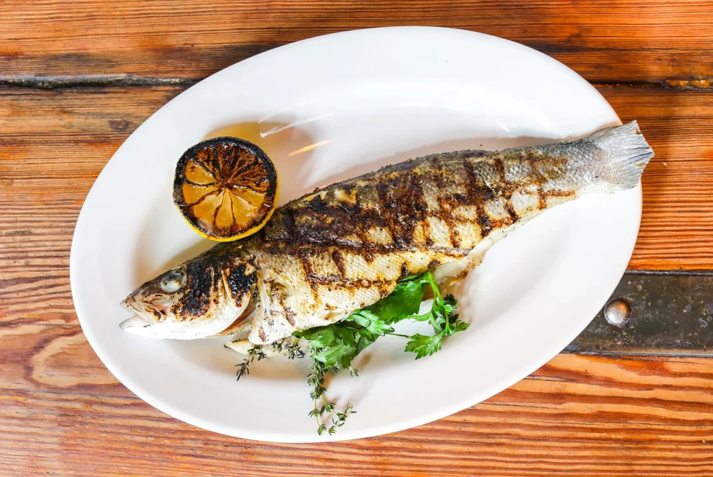 Grilled whole fish on a white plate with charred lemon and herbs.