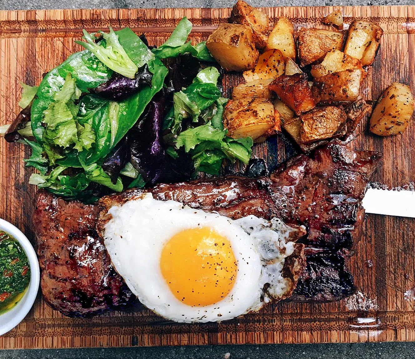 A steak, fried egg, roasted potatoes, and salad on a wooden board.