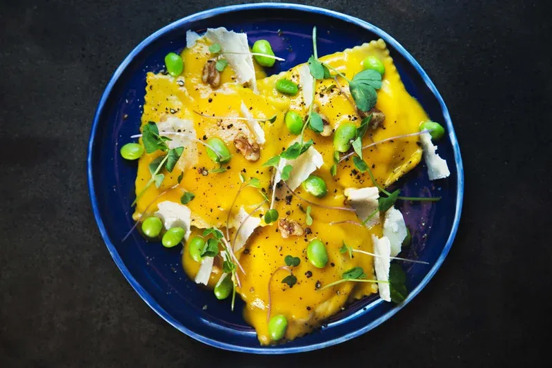 Yellow ravioli with green beans, walnuts, cheese, and microgreens on a blue plate.