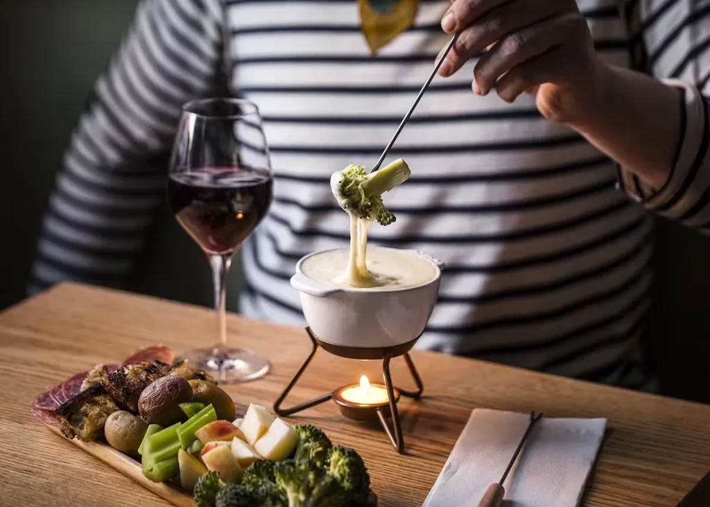 A person dips broccoli into a fondue pot, with wine and sides.