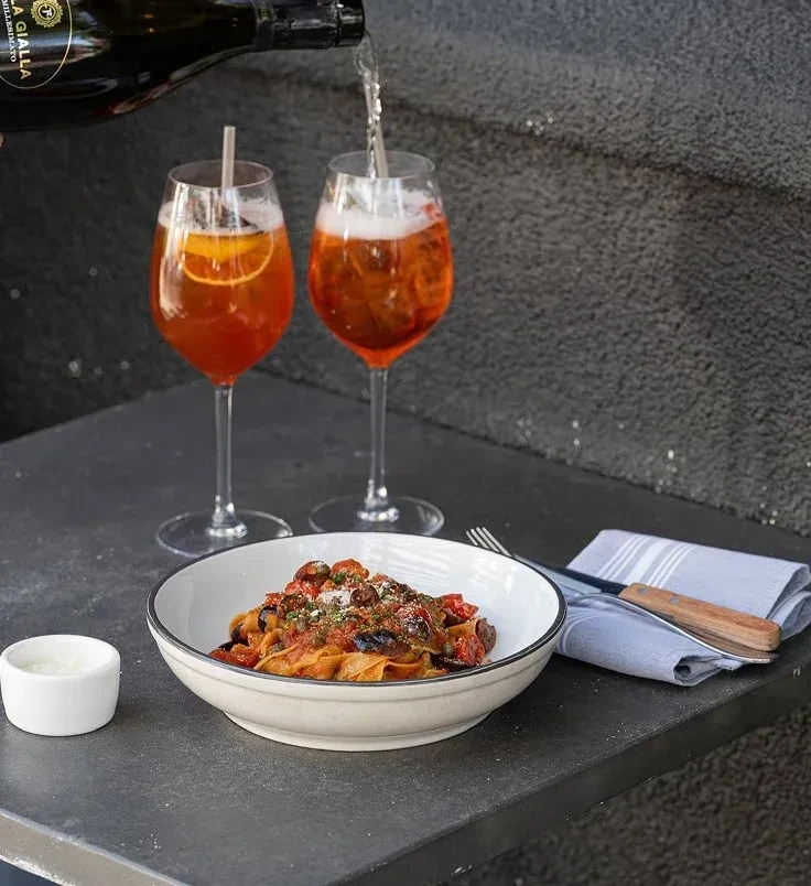 Pouring champagne into a glass next to pasta and another drink.