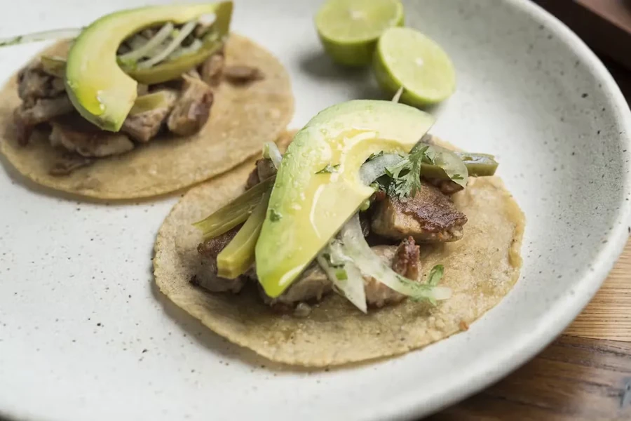 Two tacos with meat, avocado, onions, cilantro, and lime wedges.