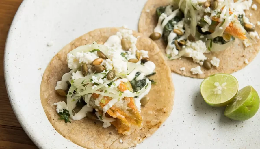 Two tacos with white cheese, pumpkin seeds, and lime on a white plate.