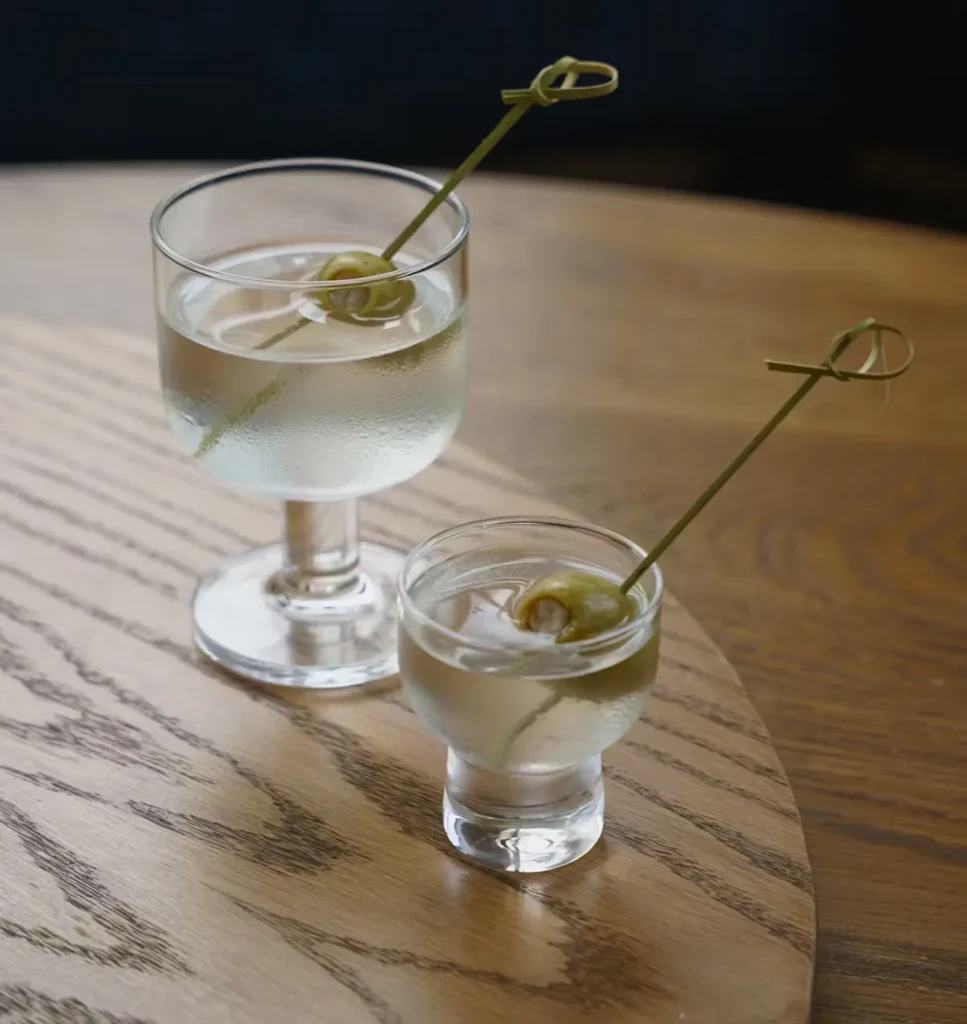 Two clear glasses of gin with olive garnishes on a wooden surface.