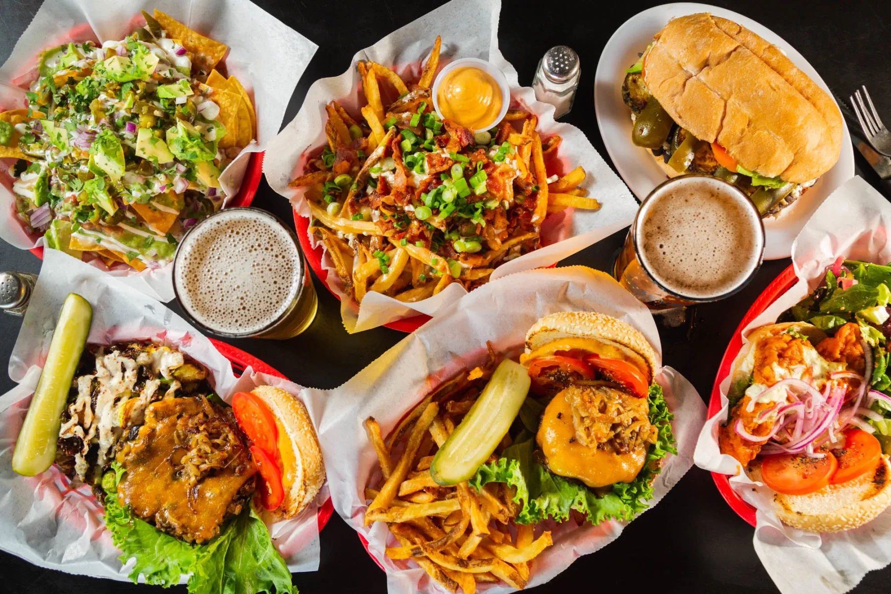 Overhead view of several meals and beers on a dark table.