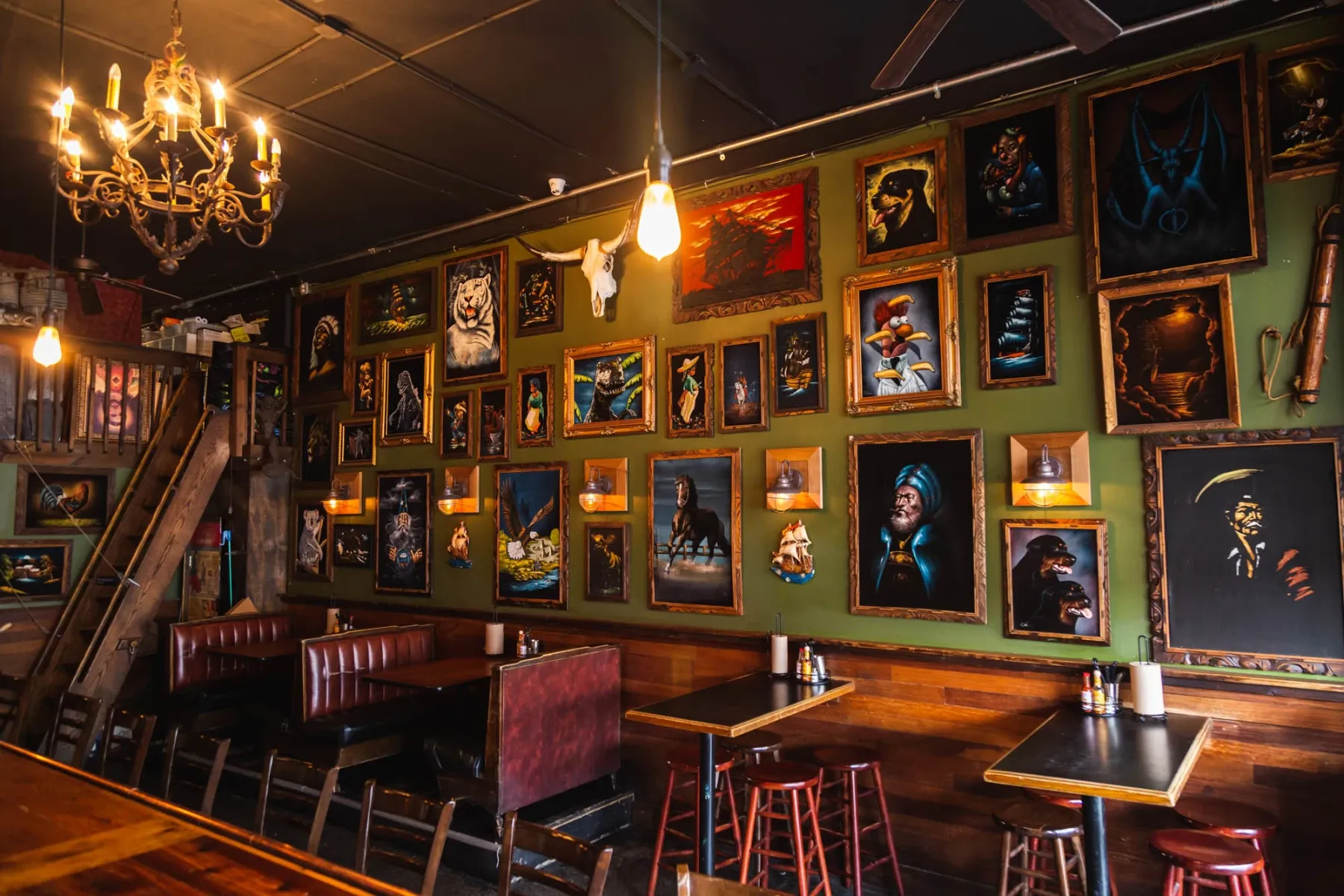 Eclectic bar interior with framed art, a chandelier, and a wooden staircase.
