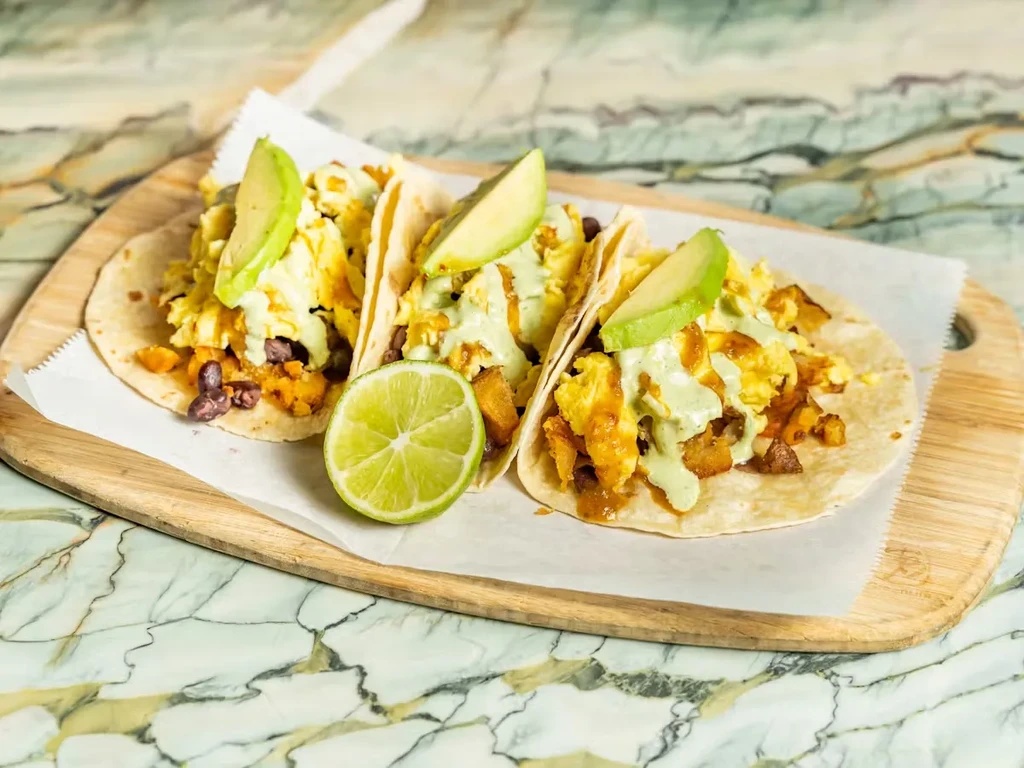 Three breakfast tacos with avocado, scrambled eggs, and lime on a board.