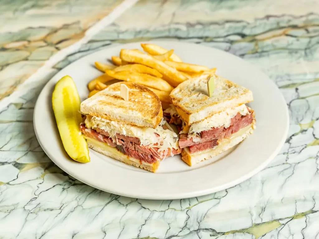 Reuben sandwich, French fries, and pickle spear on a marble surface.
