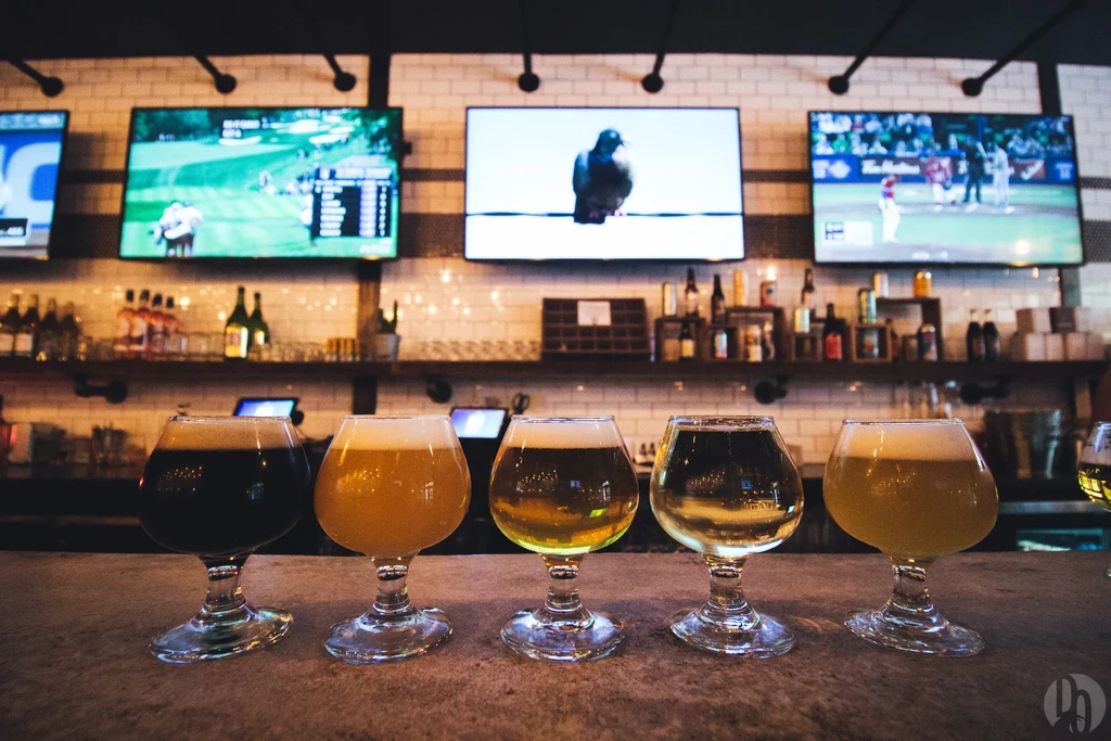 Five glasses of beer in front of three TVs in a bar.