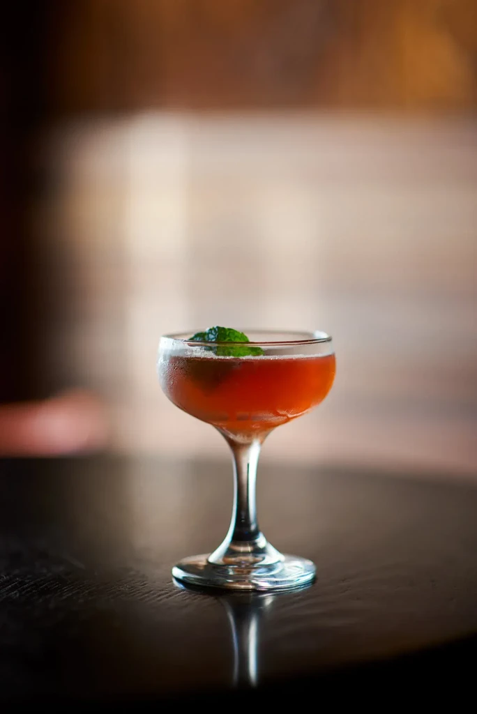 A coupe glass of red cocktail with a mint leaf garnish.