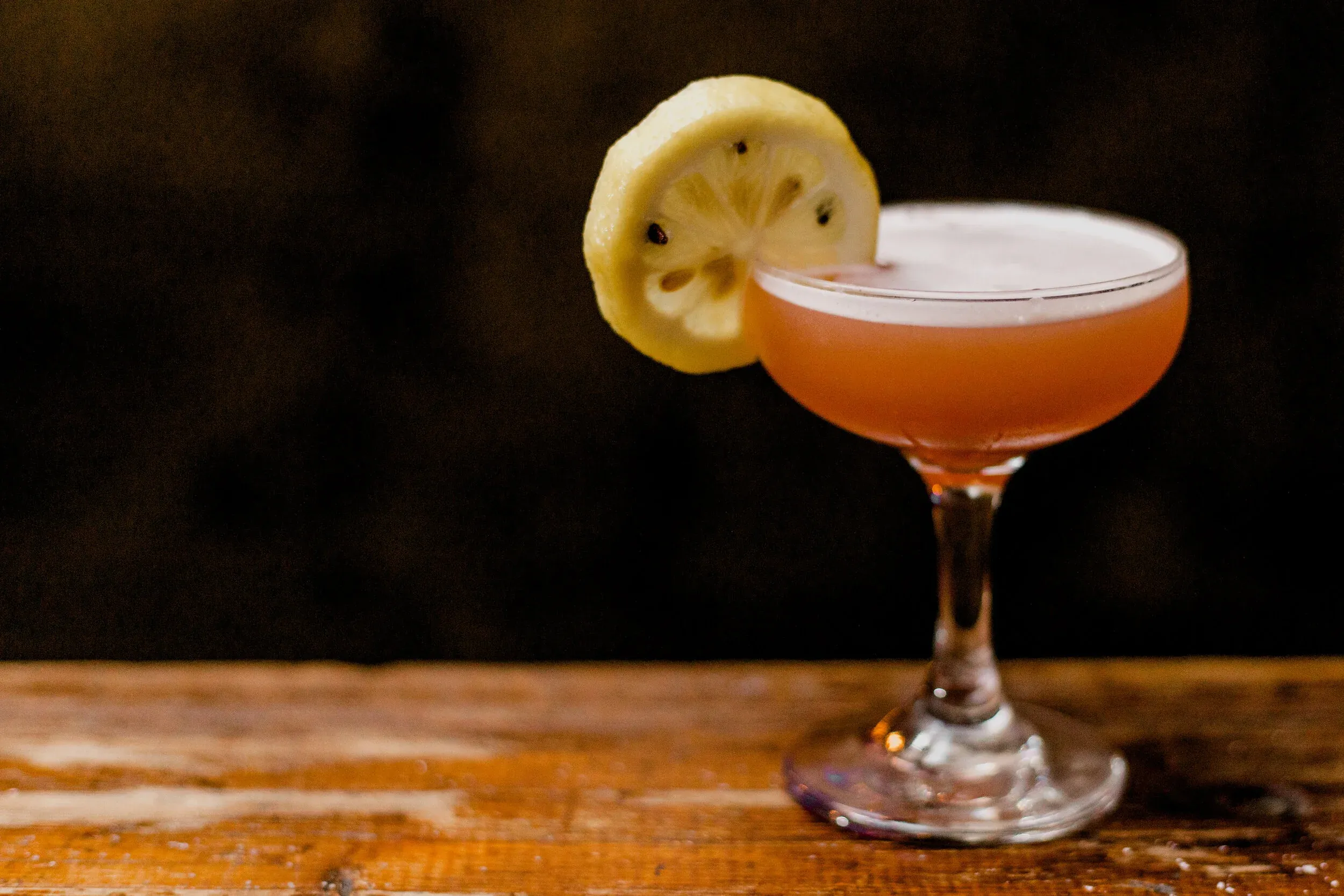 Pinkish cocktail in coupe glass with lemon slice on wooden surface.