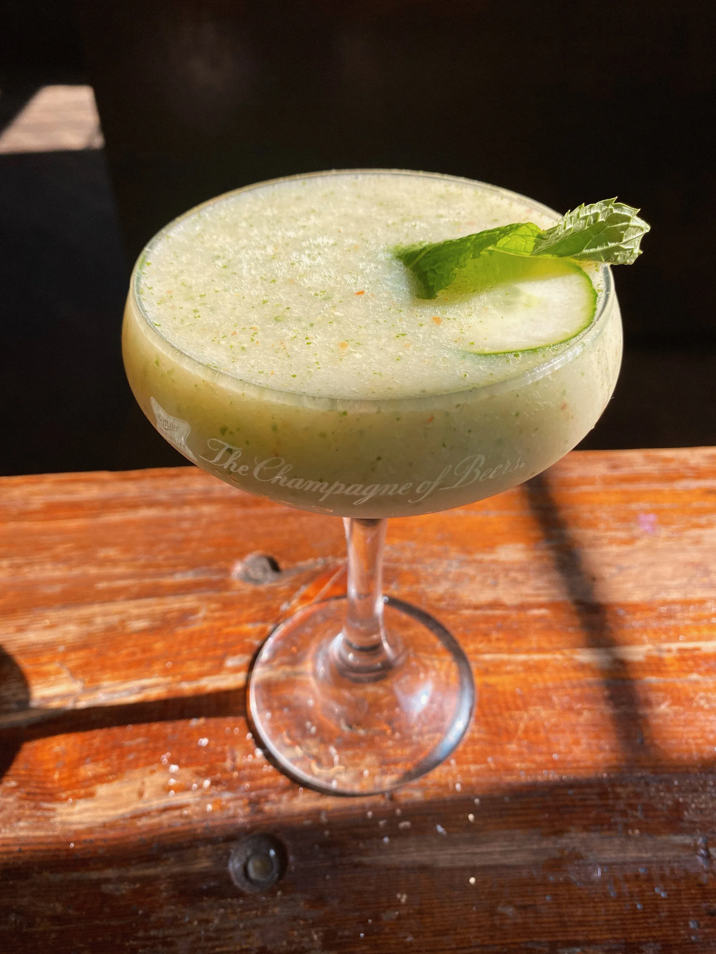 A pale green cocktail with a cucumber slice and mint leaf on a wooden surface.