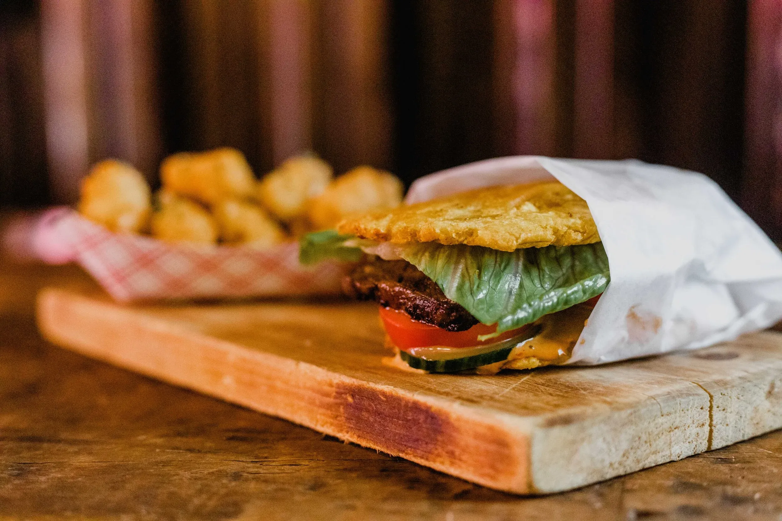 A burger wrapped in paper with fries in a basket on a wooden board.