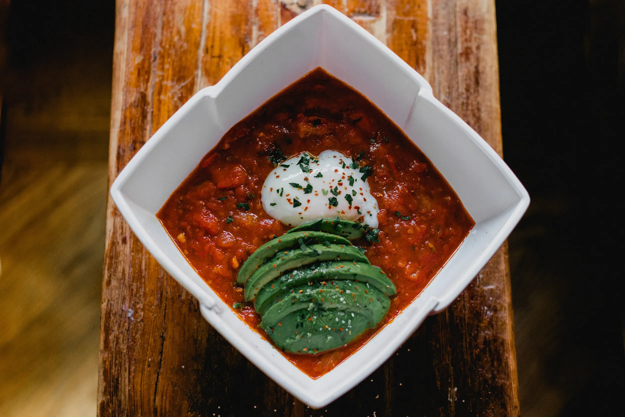 A square white dish of red stew with avocado and a poached egg.